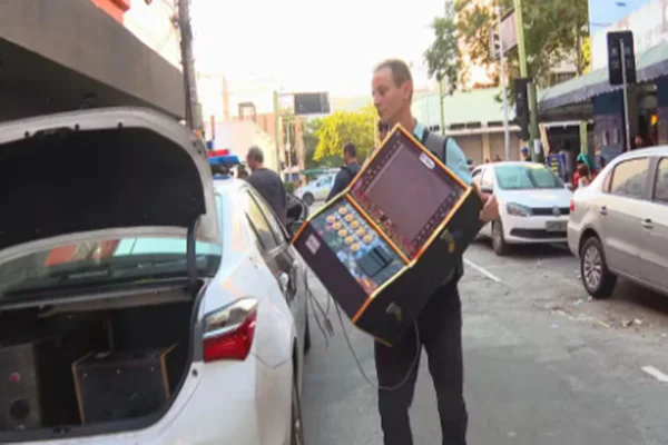 Imagem colorida de policial colocando máquina caça-níquel em porta-malas de veículo - Metrópoles