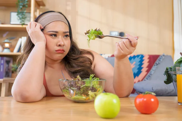 Foto colorida de mulher fazendo careta ao olhar para um garfo com salada - Metrópoles