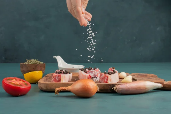 Foto colorida de uma mão colocando sal que tempera uma tipo de carne numa tábua de madeira - Metrópoles.