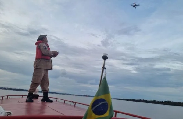 bombeiro militar está em cima de um barco que navega em um rio. ele manuseia um drone