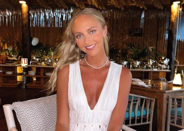 Na imagem colorida, uma mulher loira de vestido branco posa em um restaurante