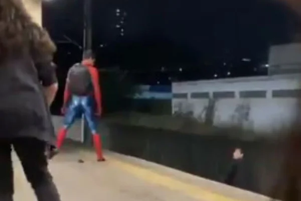 Homem vestido como personagem Homem-Aranha, roupa vermelha e azul, em plataforma de metrô, olhando para uma rapaz que está nos trilhos - Metrópoles