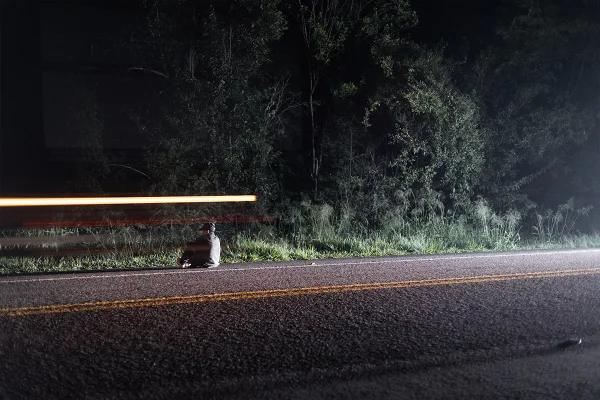 Foto mostra um jovem sentado à veira de uma estrada de costas
