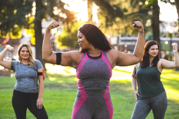 Foto colorida de trê smulheres fazendo sinal de força com os braços, vestidas com trajes de exercício físico - Metrópoles.