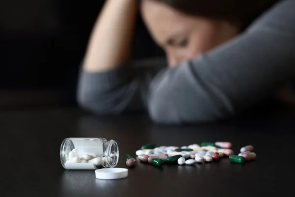 Imagem colorida: remédios antidepressivos sobre a mesa e mulher no fundo com a mão na cabeça - Metrópoles - antidepressivos