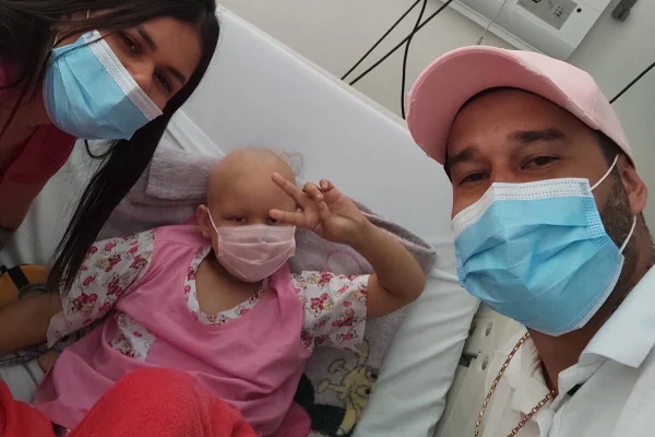 Menina careca ladeada por homem e mulher, todos com máscaras cirúrgicas, em ambiente hospitalar - Metrópoles