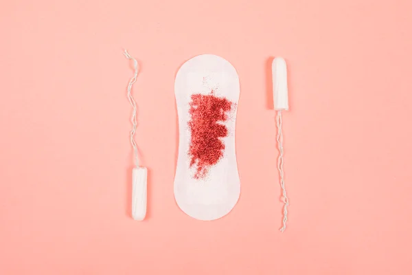 Foto mostra absorventes menstruais, um deles com sangue, em fundo rosa claro. Metrópoles