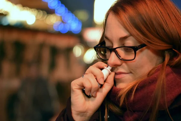 Imagem mostra mulher ruiva, de óculos, colocando um descongestionante nasal no nariz - Metrópoles