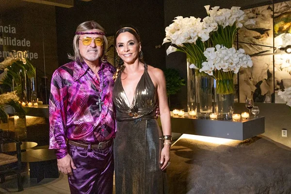 Paulo está vestido com camisa e calça roxa estilo anos 70 e com peruca branca. Ao seu lado, a esposa Denise Zuba veste um vestido longo dourado - Metrópoles