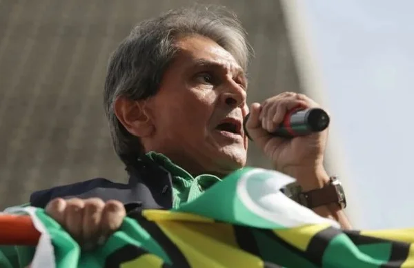 Na imagem colorida, um homem está posicionado no centro. Ele tem cabelos curtos, pele clara e olha seriamente para a frente