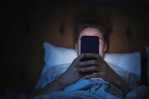Foto colorida de homem mexendo no celular deitado na cama. - nomofobia