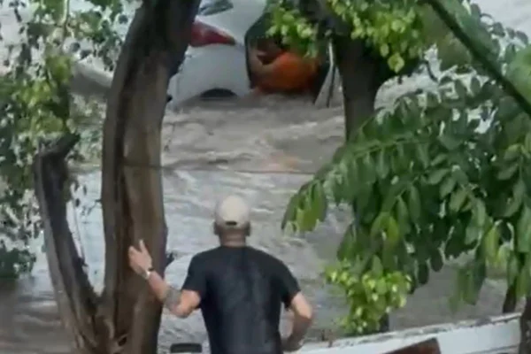 Imagens coloridas mostram carro branco sendo levado por enxurrada com uma pessoa, de camiseta laranja e cabelos brancos, dentro do veículo. Uma mulher morreu - Metrópoles