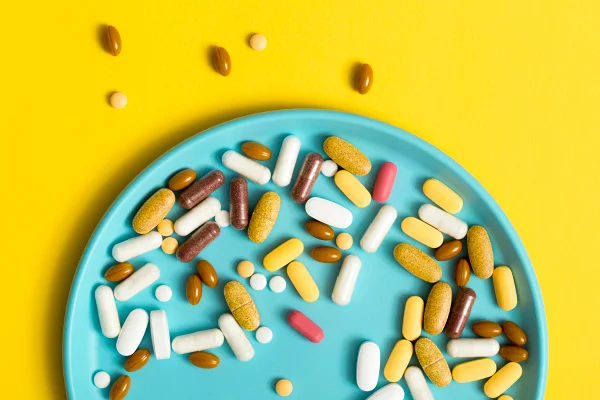 Foto colorida com fundo amarelo, de um prato cor azul, cheio de pílulas que simbolizam suplementos alimentares - Metrópoles