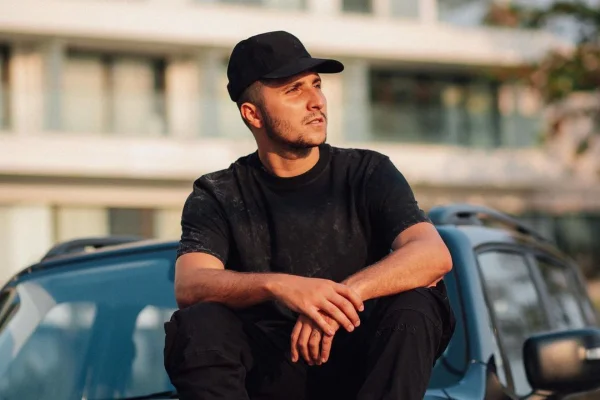 Foto colorida de Matheus Novinho usando boné e roupa pretas, sentado em carro - Metrópoles