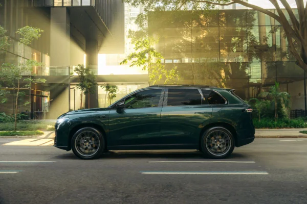 SUV verde metálico está estacionado em frente a um edifício moderno de vidro e concreto, sob a luz amarelada do entardecer. Árvores refletem na lataria do carro.