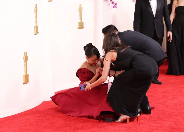 Liza Koshy sendo ajudada por duas pessoas após tropeçar e cair no tapete vermelho do Oscar 2024