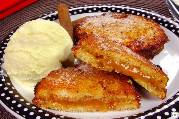 Na foto temos um prato branco de bordas pretas com bolinhas brancas e dentro rabanadas recheadas com creme marrom claro e partidas ao meio e uma bolsa de sorvete amarelo claro ao lado