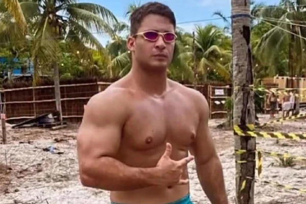 Foto colorida de homem branco, sem camisa, de óculos escuros e em quadra de areia