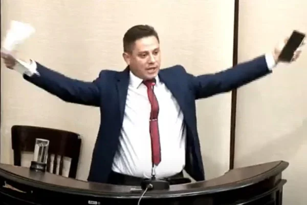 Homem branco vestido com "terno e gravata" fala na tribuna de Câmara de Vereadores.. Ele está com os braços abertos, tem um celular em uma das mãos e papéis em outra.