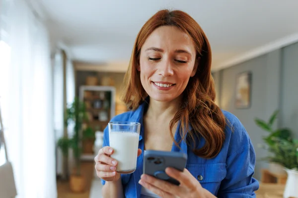 Foto colorida de mulher com blusa azul segurando um copo de leite e teclando no celular - Metrópoles
