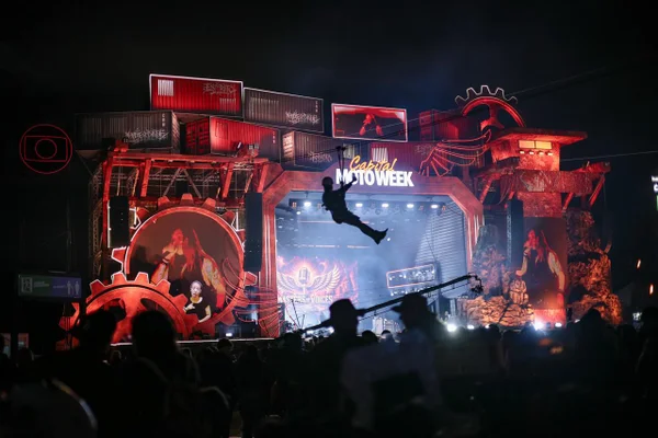 Vista do palco principal iluminado do Capital Moto Week, com estrutura de engrenagens e contêineres, tirolesa em ação e público na Cidade da Moto, em Brasília.