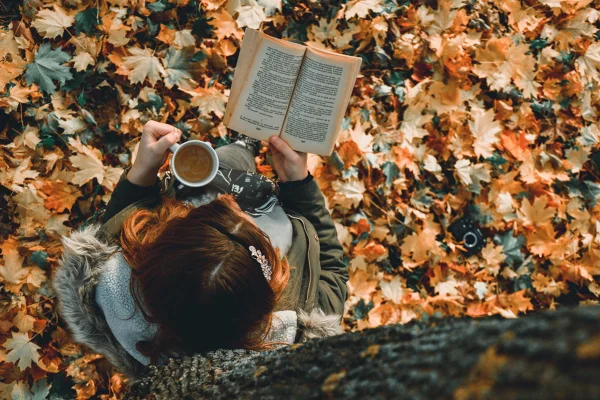 Imagem colorida de mulher lendo livro e tomando chá sentada no chão rodeado de folhas - Metrópoles