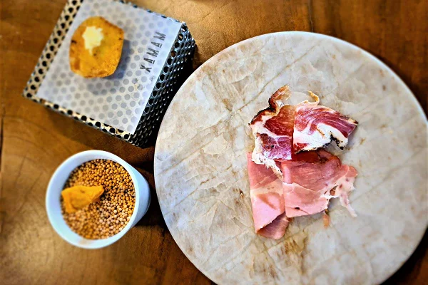 Na foto temos uma mesa de madeira com uma placa redonda de marmore branco usada como prato para charcutaria e ao lado um copo com algo amarelo e um suporte com um petisco