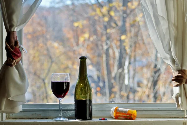 Imagem mostra uma garrafa de vinho e uma taça com a bebida ao lado de um frasco de medicamentos em uma janela. Metrópoles