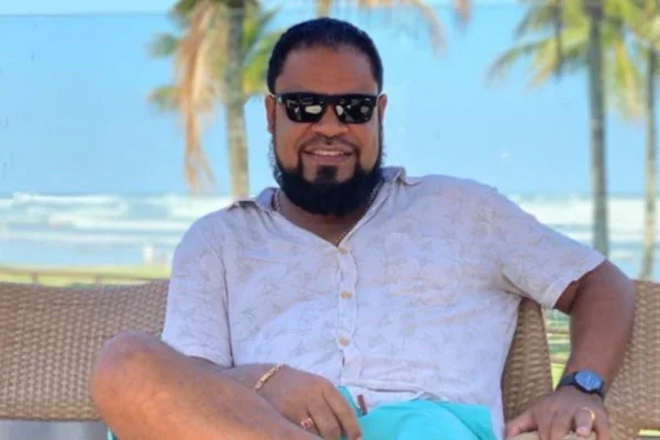 Imagem colorida mostra Latrell Brito, homem pardo, de barba preta, usando óculos escuros, camisa branca e bermuda azul, sorrindo em uma praia - Metrópoles
