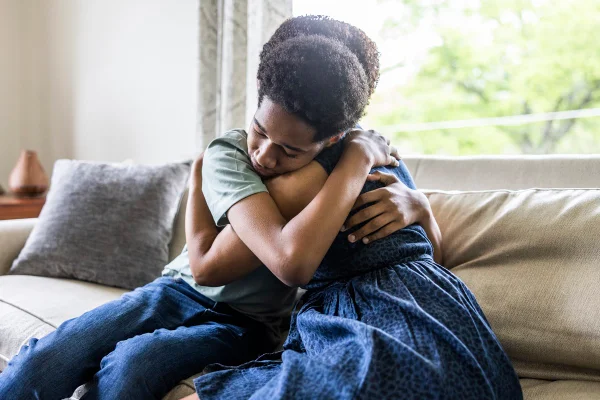 Imagem mostra adolescente abraçando a mãe. Os dois estão sentados em um sofá - Metrópoles