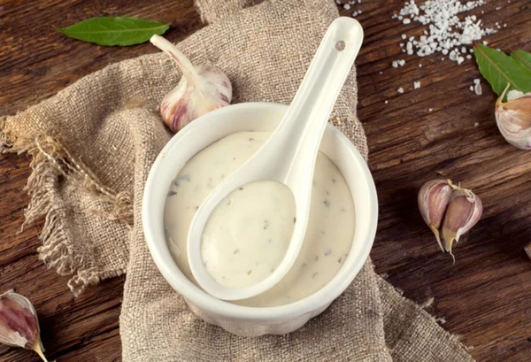 Molho aioli em um recipiente branco por cima de um pedaço de pano com dentes de alhos soltos em volta - Metrópoles