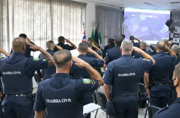 Em foto colorida batalhão de GCMs, de costas, batem continência dentro de sala de reunião - Metrópoles