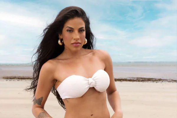 Na imagem colorida, mulher usa biquíni branco e posa na praia, com cabelos ao vento - Metrópoles