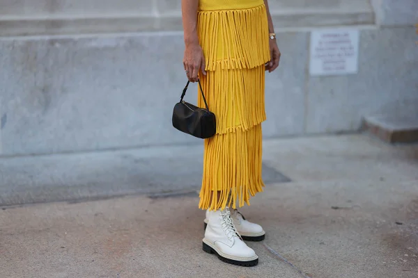 Detalhes da roupa de uma mulher durante a Semana de Moda de Nova Yrok. Na imagem, é possível ver apenas da sua cintura até os pés. Ela usa um vestido amarelo com a parte da "saia" feita de franjas; um coturno de couro branco e uma bolsa preta pequena de couro. - Metrópoles