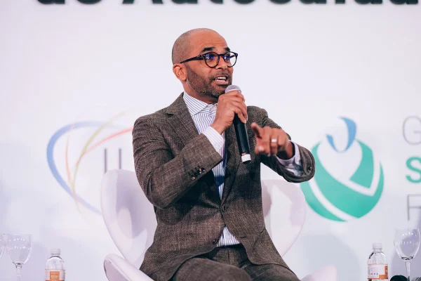 Imagem mostra Manoj Raghunandanan, um homem careca de óculos e terno, falando com um microfone na mão - Metrópoles