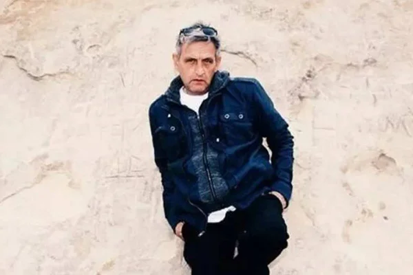 O músico Paul Ryder, que morreu aos 58 anos pouco antes de tocar em show na Inglaterra, aparece em foto posando com jaqueta e calça jeans escorado em parede branca - Metrópoles