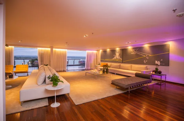 Suíte presidencial do Royal Tulip Brasília. Na foto, uma ampla sala de estar com tapete e sofás na cor clara, mesa de centro de vidro, um quadro na parede com a arquitetura de Oscar Niemayer. Ao fundo porta aberta para o deck com vista para o lago - Metropoles