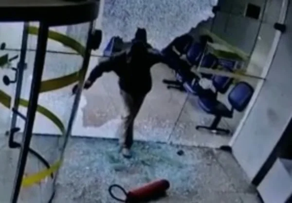 Homem de capuz entrando por porta quebrada de agência