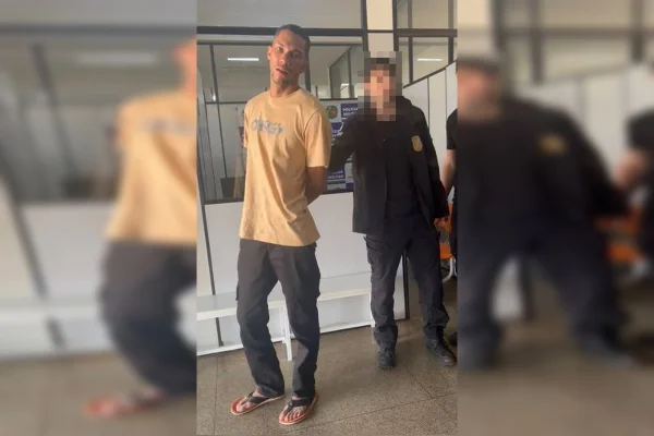 Imagem de um homem de camiseta amarela, algemado, sendo seguro por um policial de preto