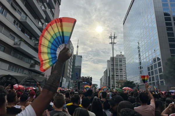 Imagem colorida mostra braço em primeiro plano com leque com as cores do arco-íris na mão direita. Ao fundo, aparece uma multidão e prédios da avenida Paulista - Metrópoles