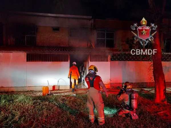 Foto colorida tirada à noite de casa iluminada por luz externa vermelha e homem com uniforme de bombeiro o