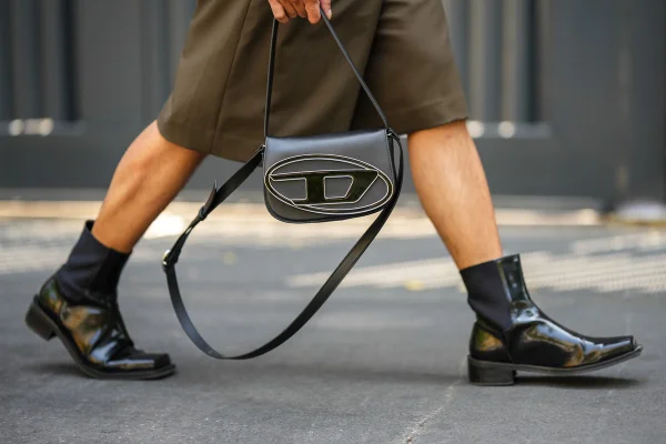 Pessoa branca e jovem andando pela rua. Na imagem é possível ver apenas suas pernas e uma de suas mãos. Usa uma bermuda verde larga, uma bota preta de couro e segura a bolsa 1DR da marca Diesel, também preta e de couro. - Metrópoles