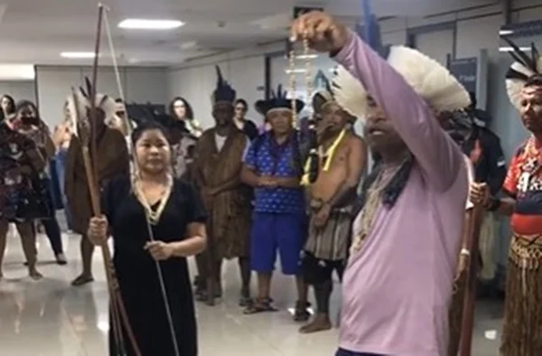 Indígenas Pataxó fazem ritual de "limpeza espiritual" no Ministério da Saúde. Na imagem, em primeiro plano, um líder segura colar e mulher ao lado segura arco e flecha enquanto outros membros da tribo, caracterizados, observam em corredor - Metrópoles