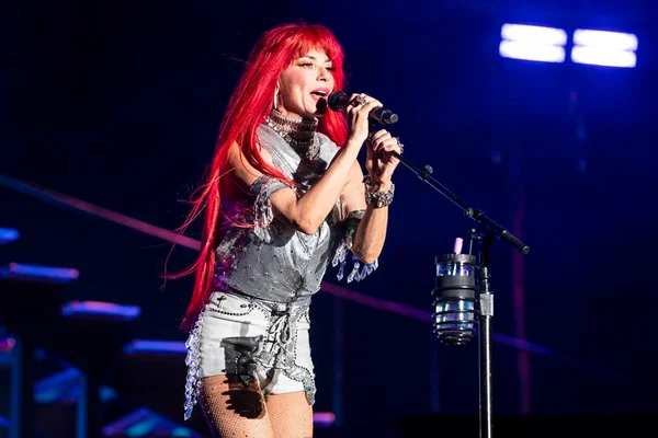 Foto colorida de Shania Twain com o cabelo vermelho, cantando em microfone e usando roupa descolada - Metrópoles
