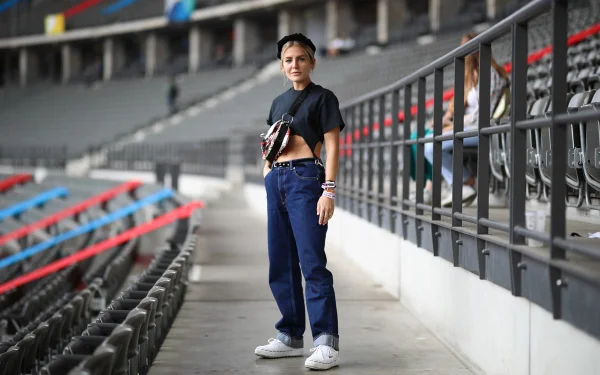 Mulher branca e jovem, de cabelo loiro amarrado em um coque, posando para foto na arquibancada de um estádio em Berlim, na Alemanha. O local é sede de shows de músicos durante o festival Lollapalooza. Ela usa uma camiseta cinza cropped, calça jeans folgada, um tênis branco e uma pochete branca de couro com calça preta.
