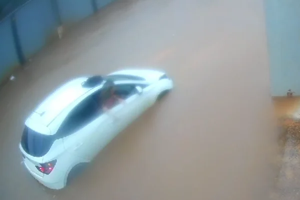 Imagem colorida de uma mulher loira, vestida com blusa rosa, descendo de um carro branco pela janela. O carro está em meio a uma enxurrada