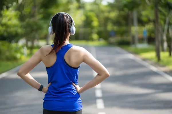 Mulher de costas com fone de ouvido e mãos na cintura em meio de exercício - Metrópoles