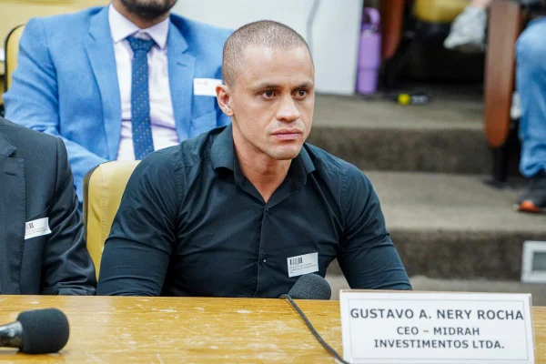 imagem colorida mostra homem com camisa preta sentado em cadeira amarela. à frente dele uma placa tem escrito o nome gustavo a nery, ceo midrah investimentos