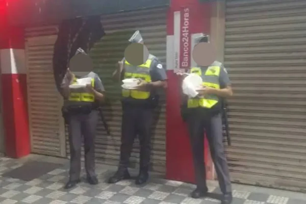 Imagem colorida de policiais militares (PMs) comendo marmitas em pé na rua - Metrópoles