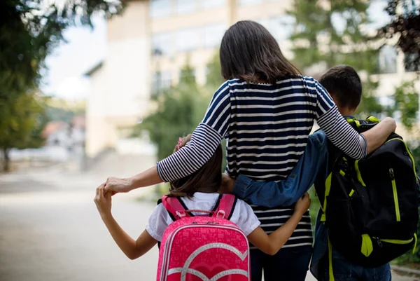 Foto colorida de mulher caminhando abraçada com duas crianças de mochila - Metrópoles